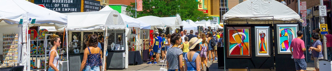 A lively scene from the Ann Arbor Art Fair, with artist booths, colorful artwork, and a crowd of visitors exploring under a sunny sky.