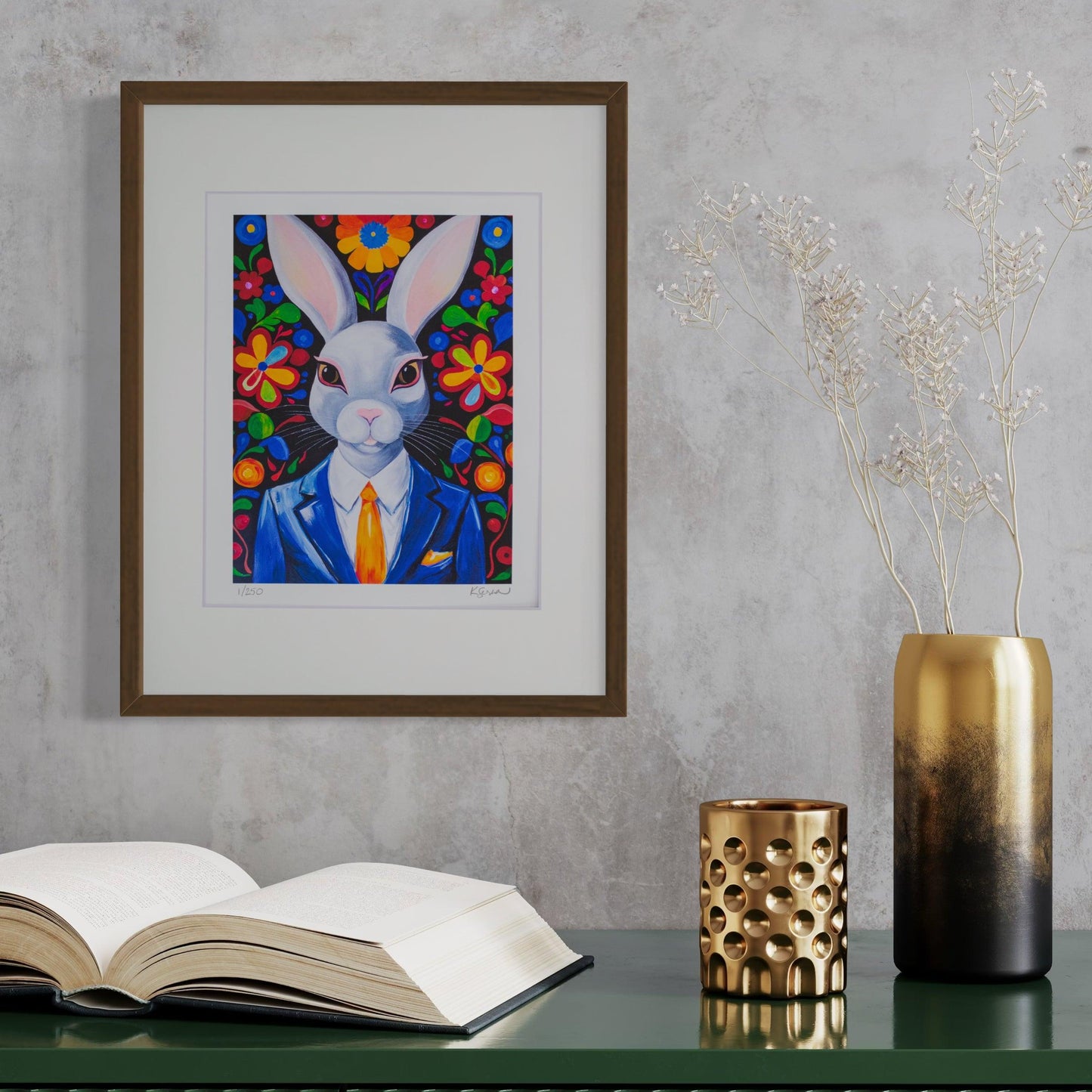 A framed print of Archie the Rabbit hangs on a textured gray wall, bringing a pop of color to the space. Archie, a regal white rabbit with striking pink eyes, wears a deep blue suit with a crisp white shirt and an orange tie. The background is filled with vibrant, oversized flowers in bright red, yellow, blue, and green. Below the artwork, a dark green tabletop holds an open book, a metallic gold vase with delicate dried flowers, and a gold perforated candle holder, creating an elegant & sophisticated scene