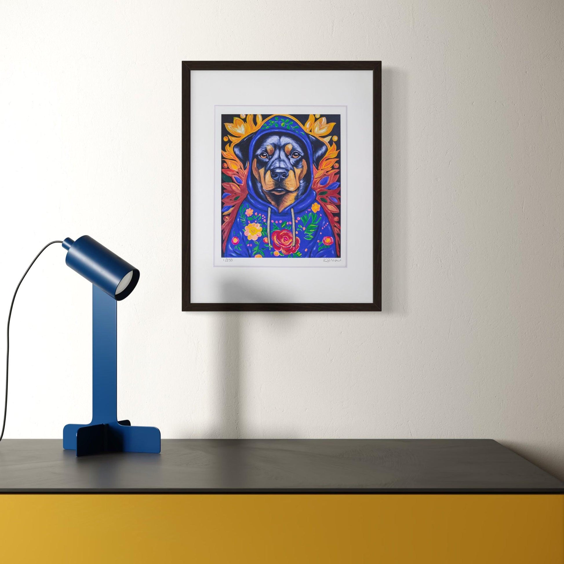 A framed print of Buster the Rottweiler hangs on a light-colored wall, adding warmth and depth to the space. Buster, a strong and expressive rottweiler, wears a deep blue hoodie covered in colorful floral embroidery, with golden autumn leaves framing his head like a crown. Below the artwork, a modern black and yellow desk is accented with a sleek blue desk lamp, giving the room a contemporary yet artistic feel.