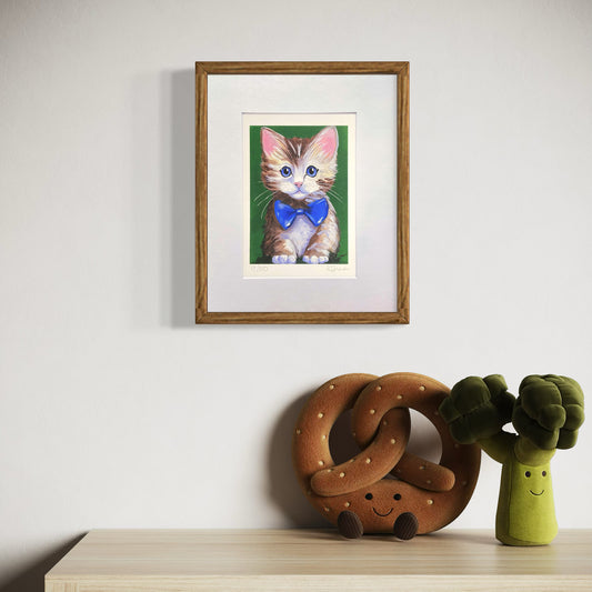 Framed artwork of a cat with a bow tie on a wall above a table with plush pretzel and broccoli toys.
