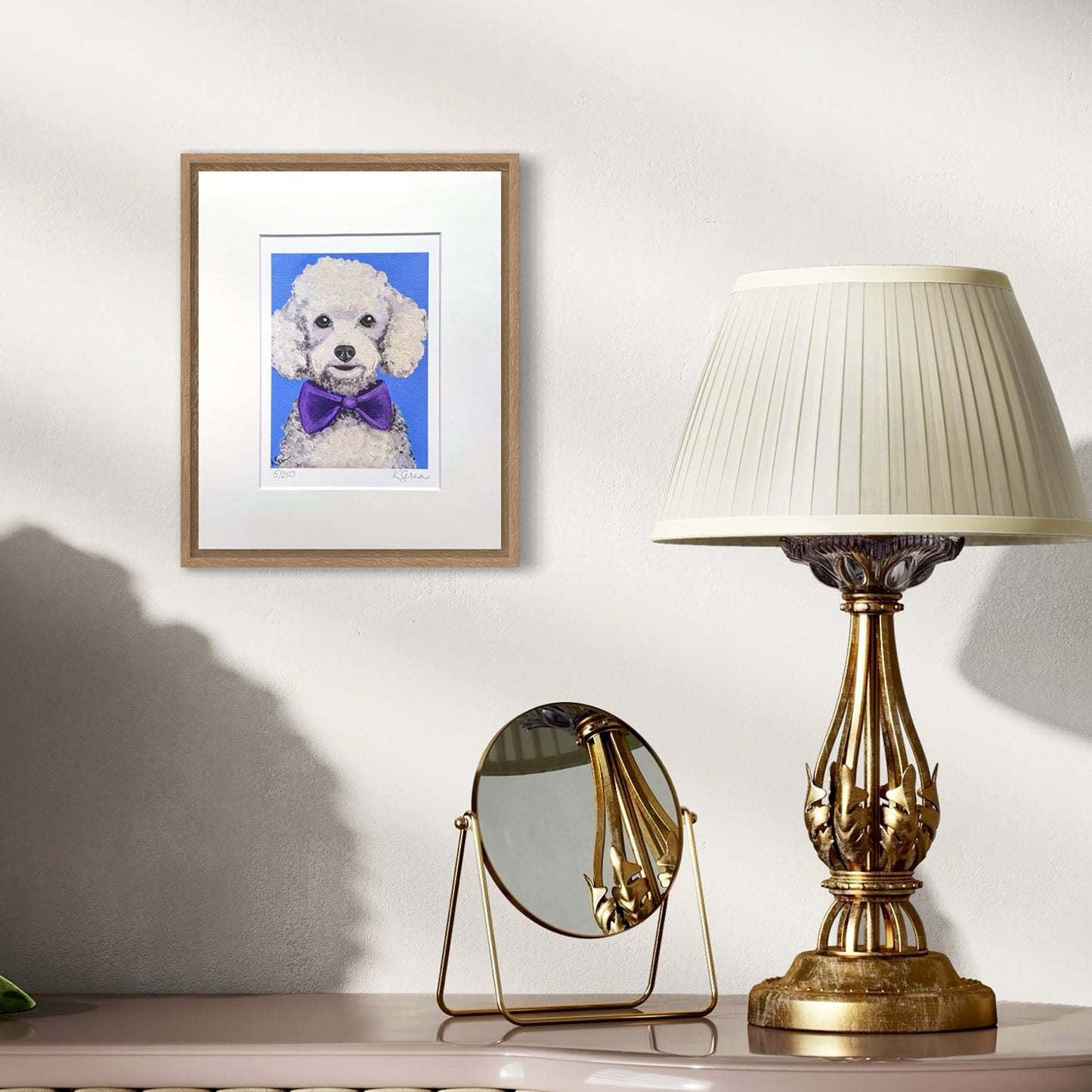 Framed artwork of a dog with a purple bow tie on a wall above a table with a lamp and mirror.