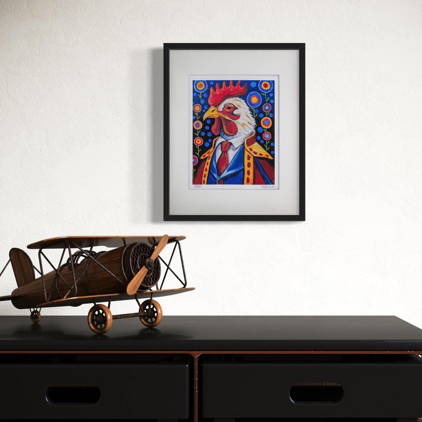 A framed print of Cornelius the Rooster hangs on a crisp white wall, adding a touch of regal charm. Below the artwork, a black console table features a vintage wooden model airplane, emphasizing an old-world, adventurous aesthetic. The bold black frame of the artwork contrasts beautifully against the clean white background, making Cornelius stand out.