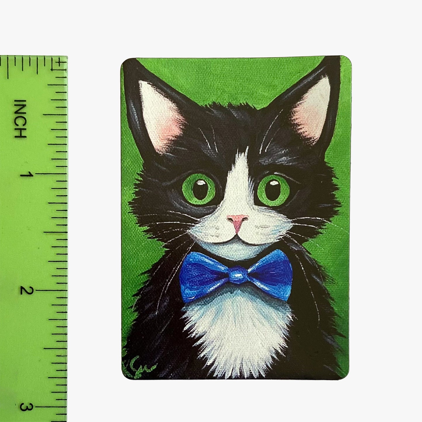 Magnet with a black and white cat wearing a blue bow tie on a green background, next to a ruler for scale.