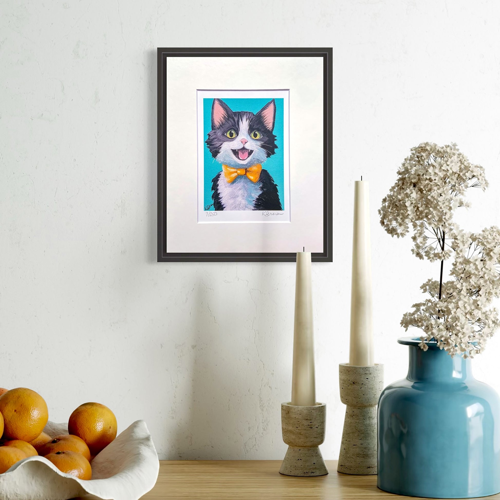 Framed artwork of a cat with a bow tie on a wall above a table with decorative items.