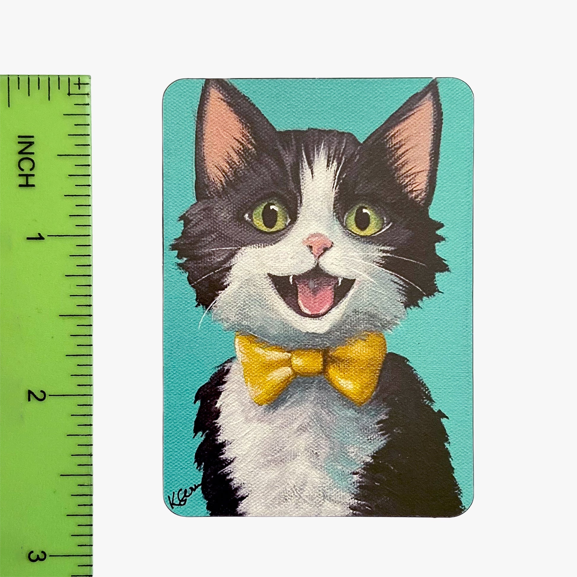 Magnet with a cartoon cat wearing a yellow bow tie on a teal background, next to a ruler for scale.