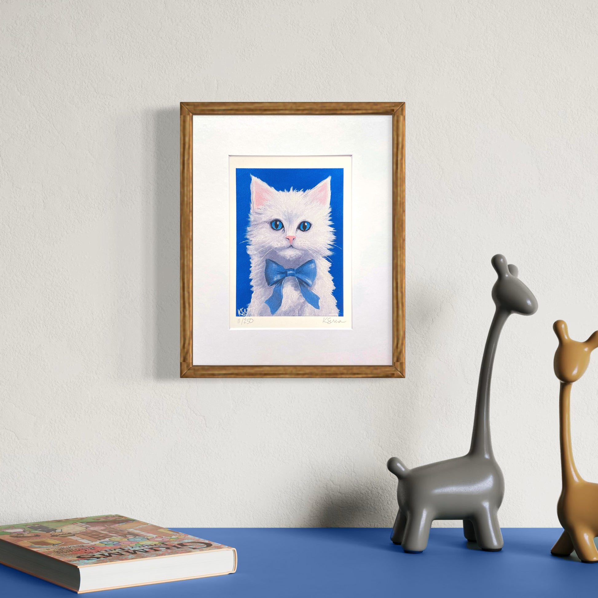 Framed artwork of a white cat with blue eyes on a wall above a blue surface with decorative items.
