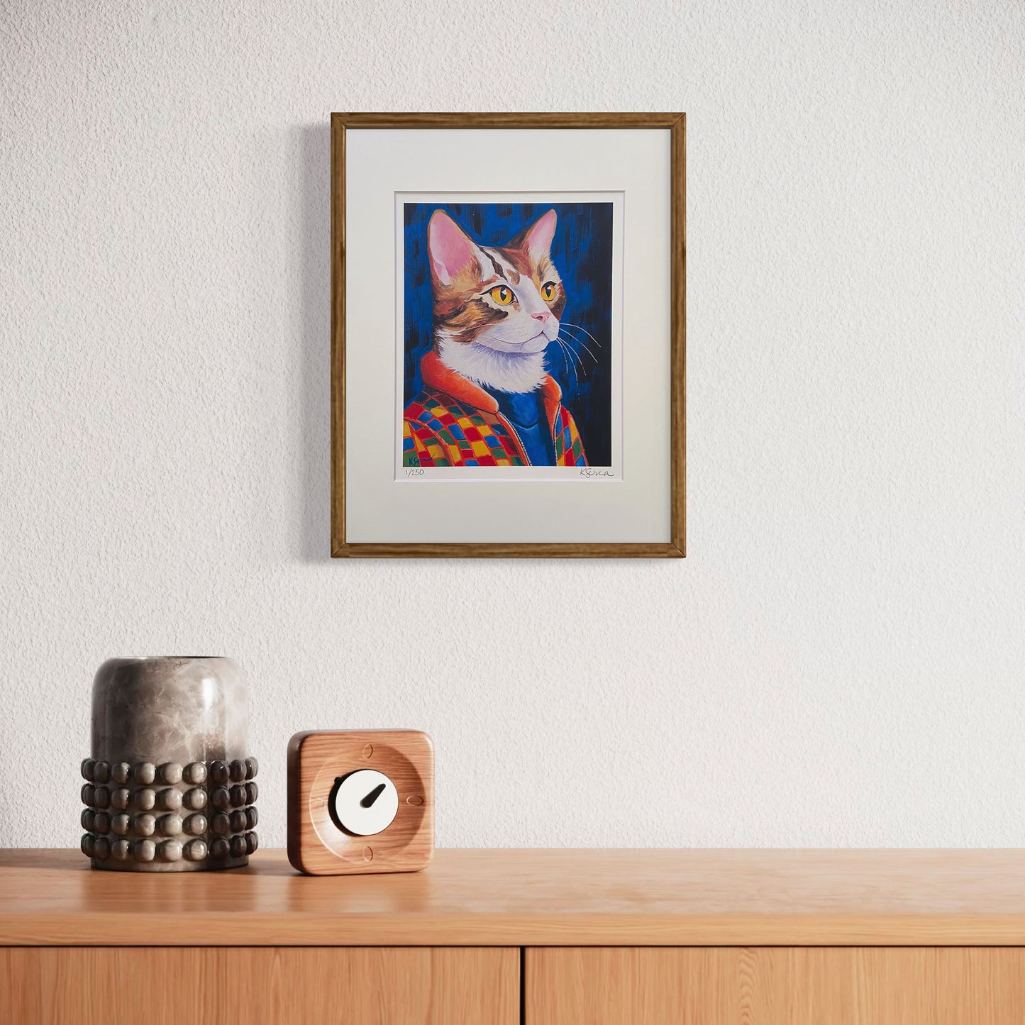 A framed print of Jasper the Cat hangs on a minimalist white wall, adding a pop of color to the space. The print is matted with a white border and enclosed in a natural wood frame. Below, a wooden console holds a sculptural ceramic vase and a small wooden clock, creating a modern, cozy aesthetic.