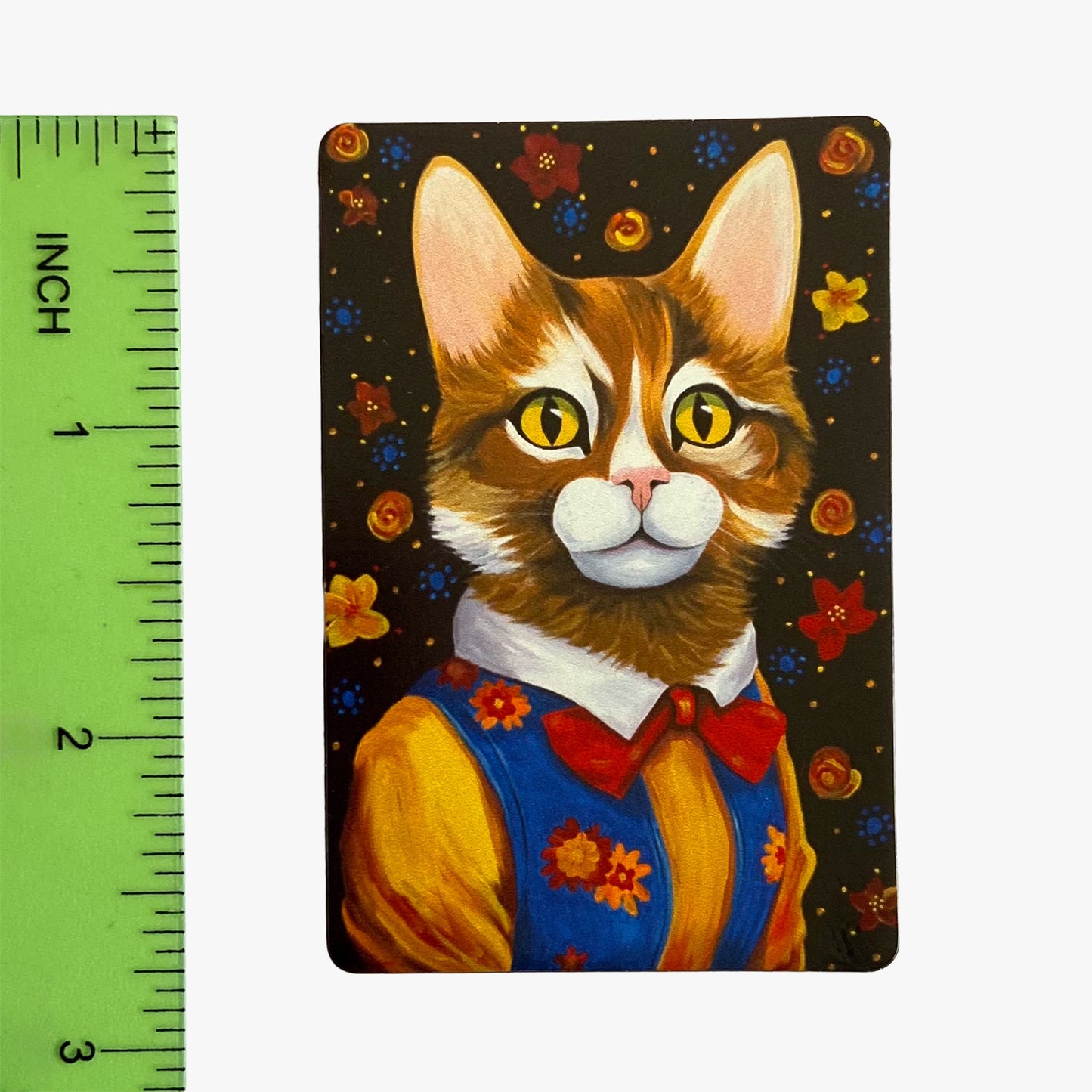 Magnet with a cat wearing a blue vest and red bow tie on a dark background with flowers, next to a ruler for scale.