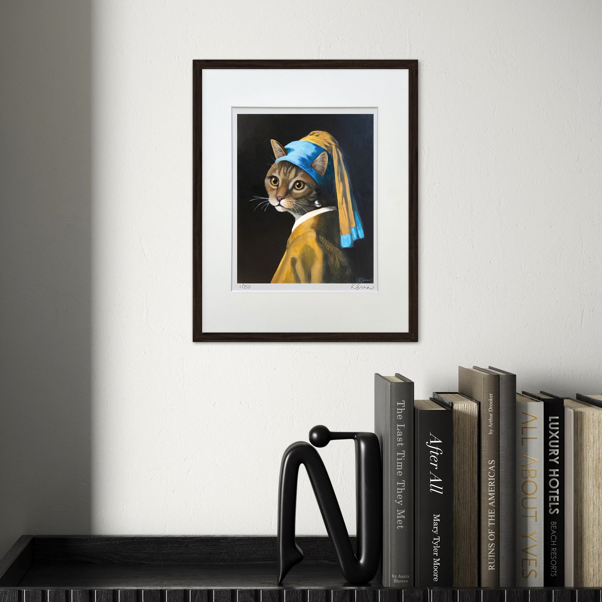 Framed artwork of a cat wearing a pearl earring on a wall above books.