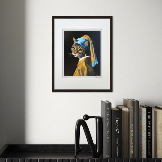 Framed artwork of a cat wearing a pearl earring on a wall above books.