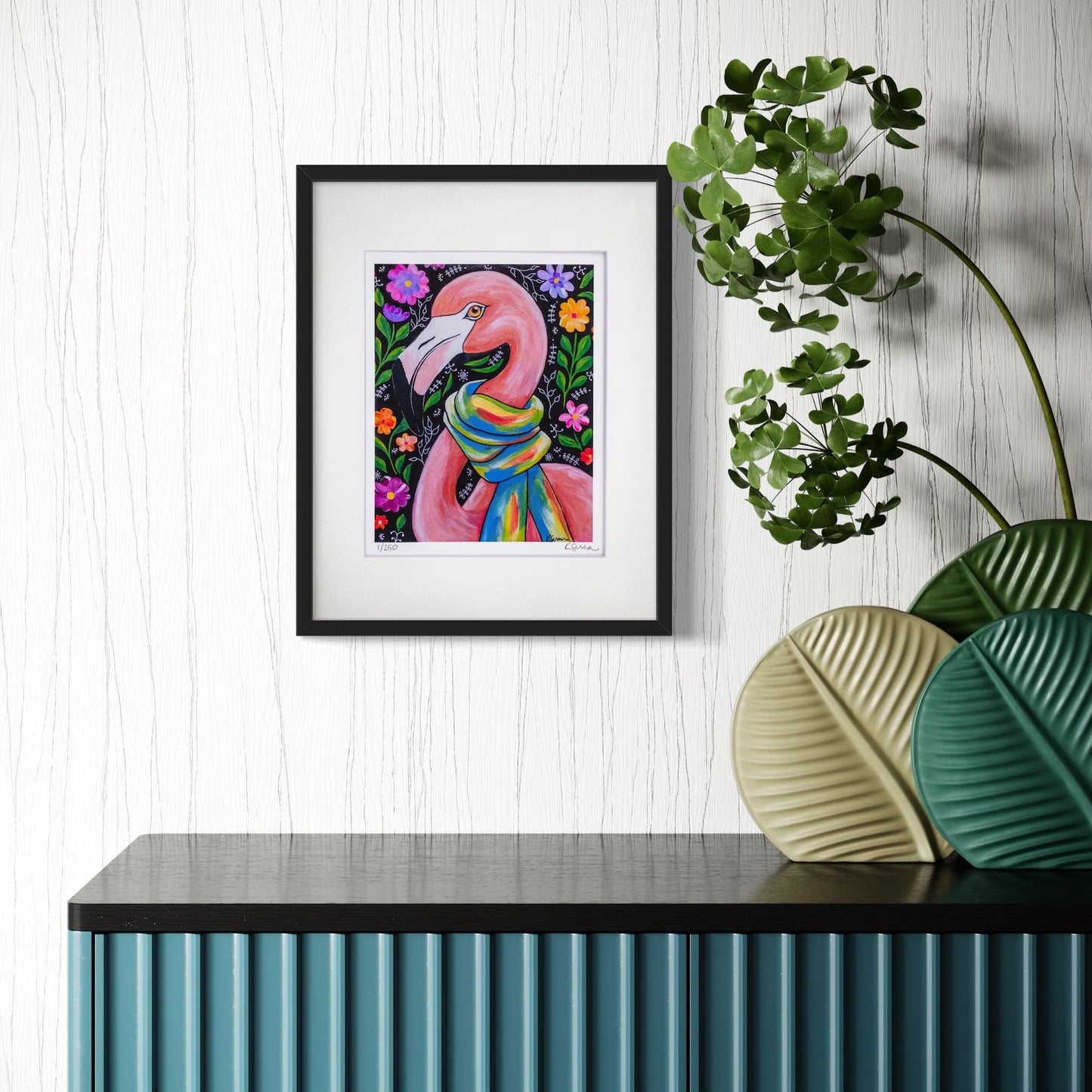 A framed print of Penelope the Flamingo hangs on a textured white wall, adding a pop of color and personality. The black frame contrasts beautifully with the vibrant artwork. Below, a modern blue console table with vertical ridges is adorned with green and beige decorative plates and a lush leafy plant, creating a fresh and lively atmosphere.