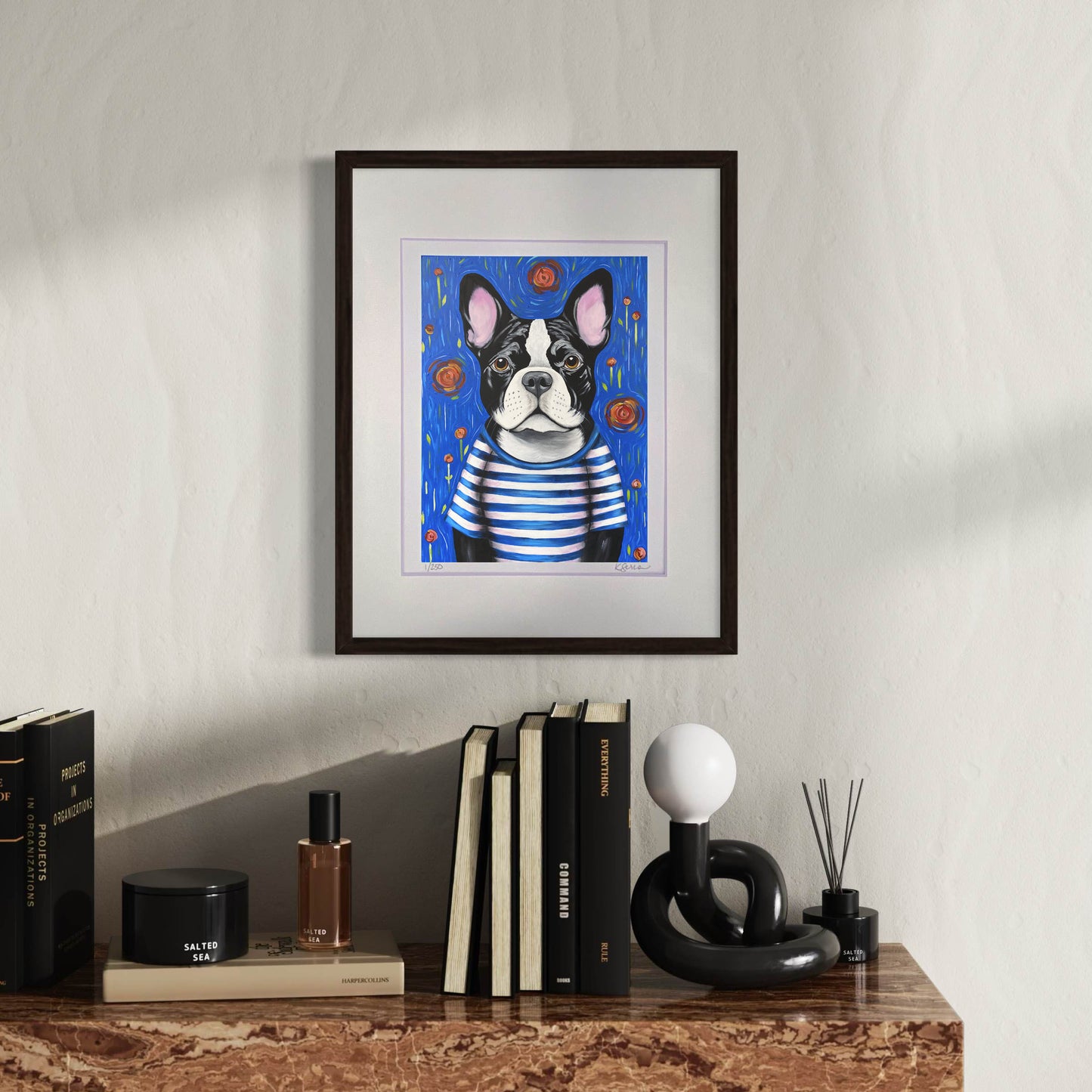 A framed print of Pierre the French Bulldog hangs on a warm-toned wall, adding a charming and playful touch. Pierre, a serious yet endearing black and white Frenchie, wears a classic striped blue and white shirt, evoking a Parisian-inspired style. The background is deep blue, dotted with expressive red and orange flowers. Below, a sophisticated wooden console holds an array of stylish black books, a modern sculpture, and a decorative candle.