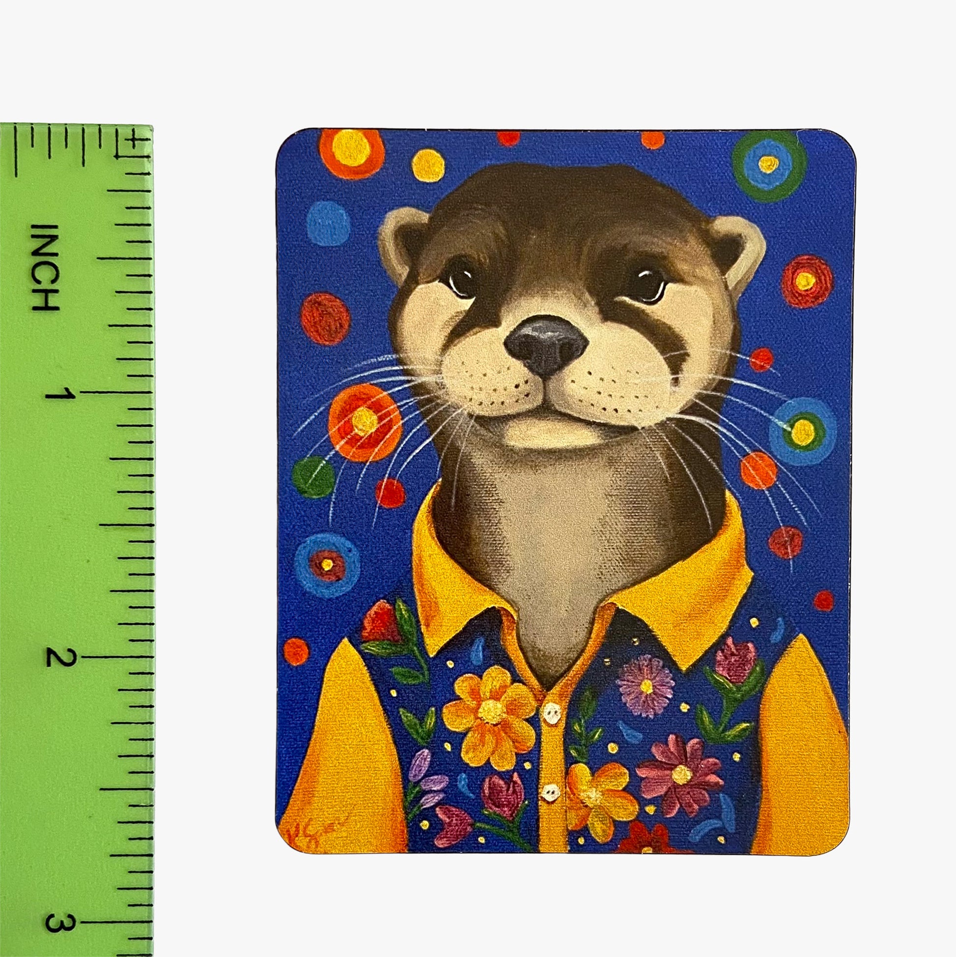 Magnet with a stylized otter wearing a yellow shirt and floral vest on a blue background with colorful flowers, next to a ruler for scale.