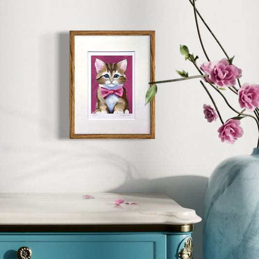 Framed artwork of a kitten with a pink bow tie on a white wall next to a blue cabinet and vase with pink flowers.