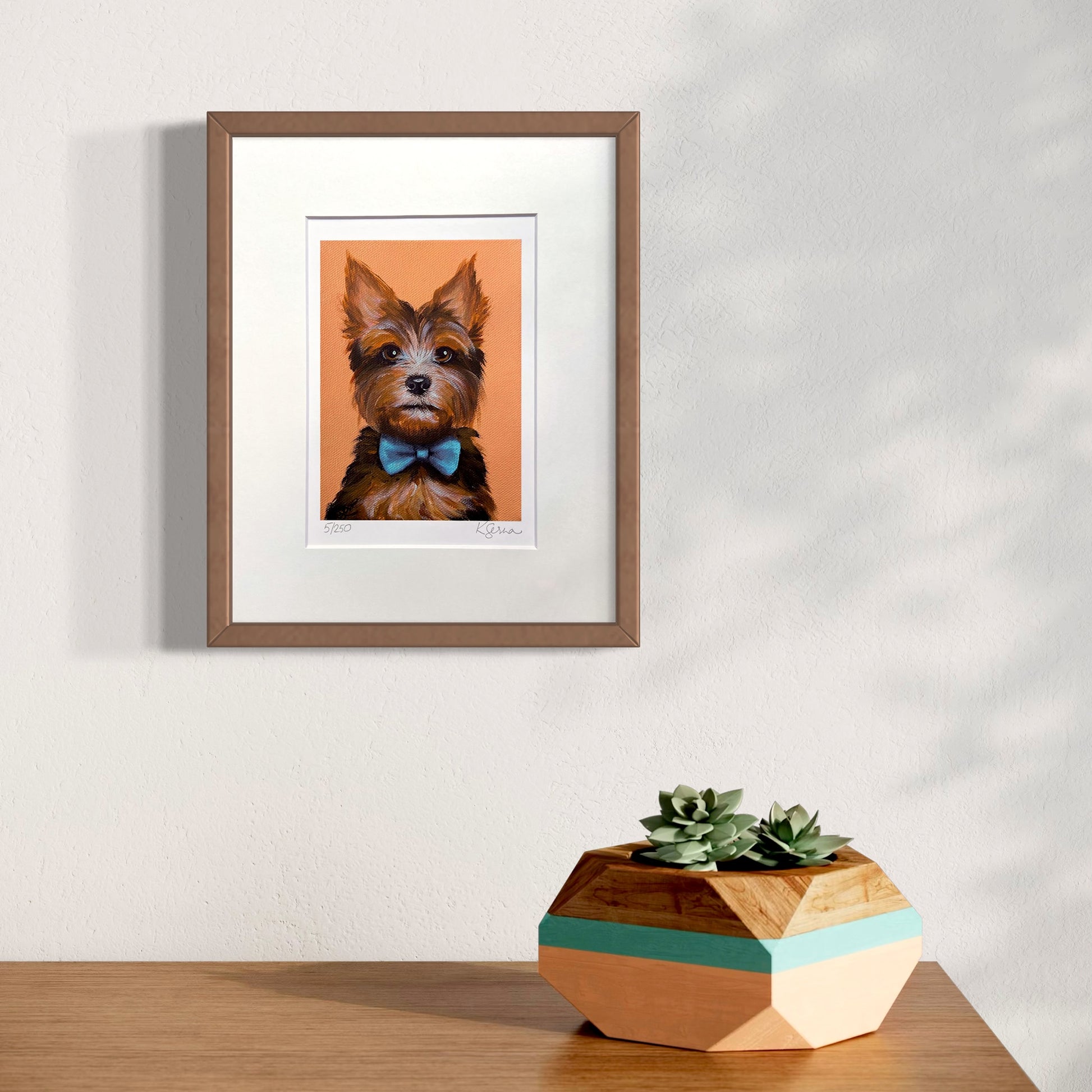 Framed artwork of a dog with a bow tie on an orange background, placed on a wooden surface with a geometric plant pot.