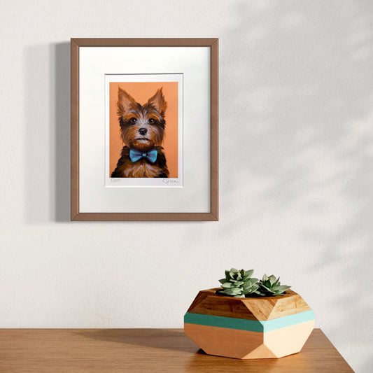 Framed artwork of a dog with a bow tie on an orange background, placed on a wooden surface with a geometric plant pot.