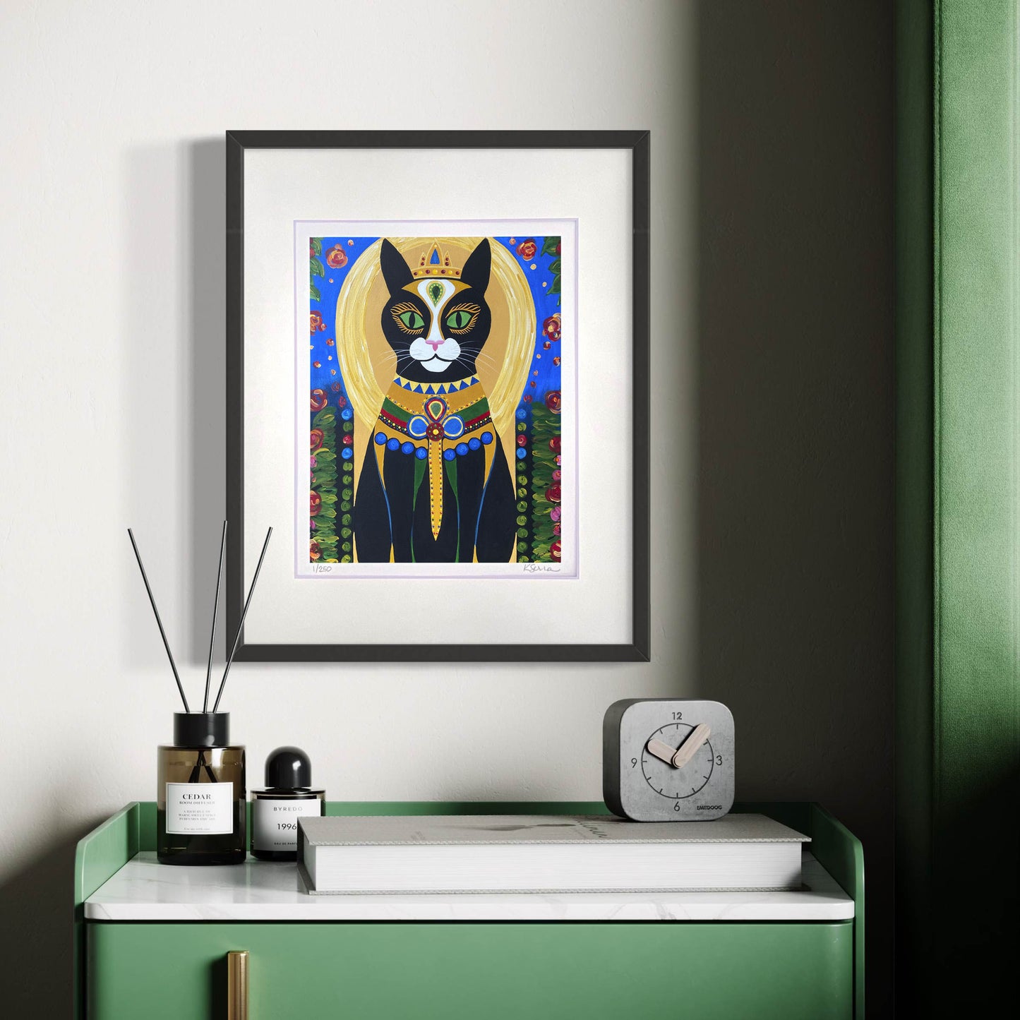A framed print of Shadow the Cat hangs on a light-colored wall, exuding a regal and mysterious presence. Shadow, a striking black cat with green eyes, is adorned in a lavish golden and blue robe decorated with intricate jewelry and an ornate collar. A golden halo and crown add to their majestic aura. Below, a sleek green and white dresser holds a minimalist clock, a candle, and a reed diffuser, creating a luxurious and elegant aesthetic.