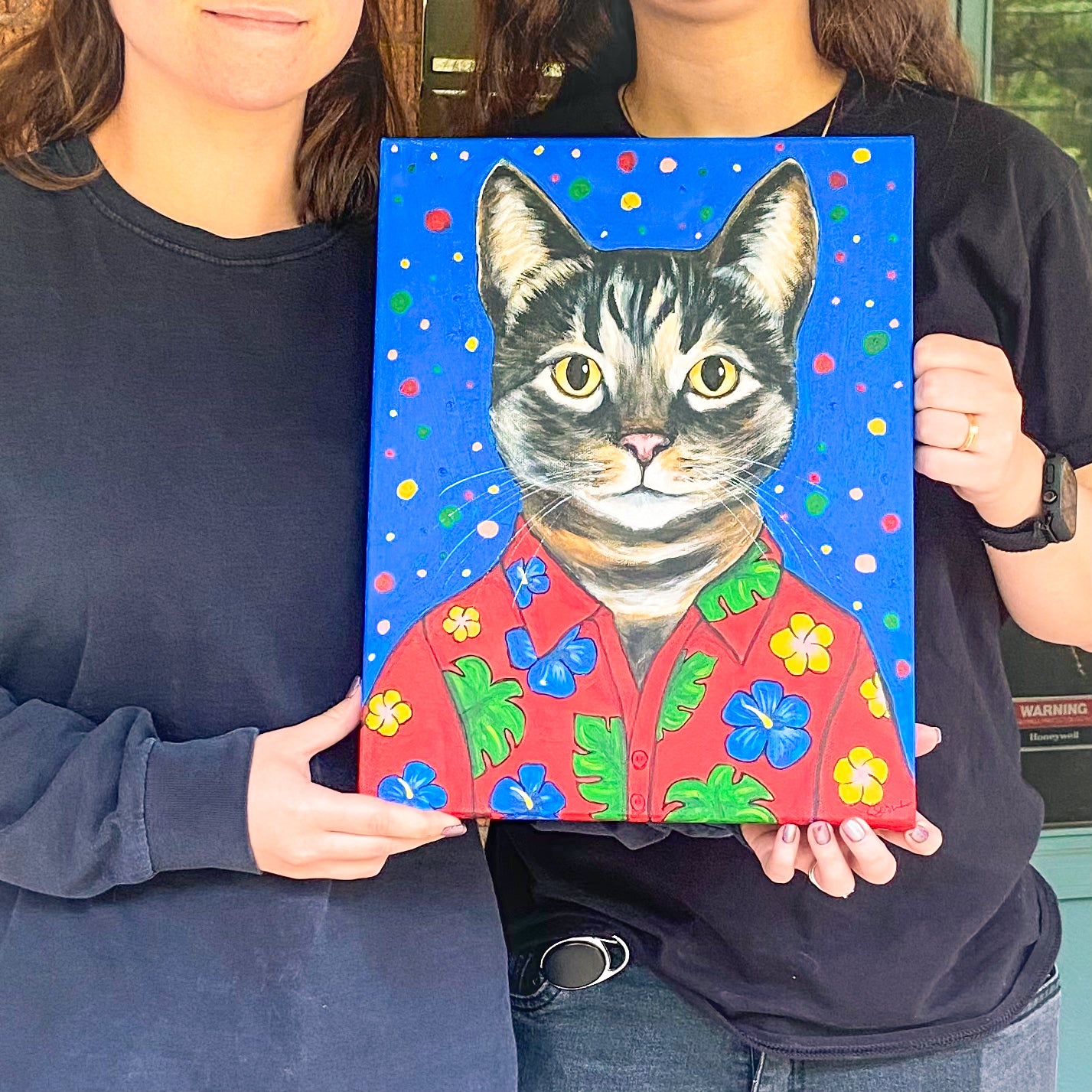 Two people holding a painting of a cat wearing a colorful shirt with a blue background.