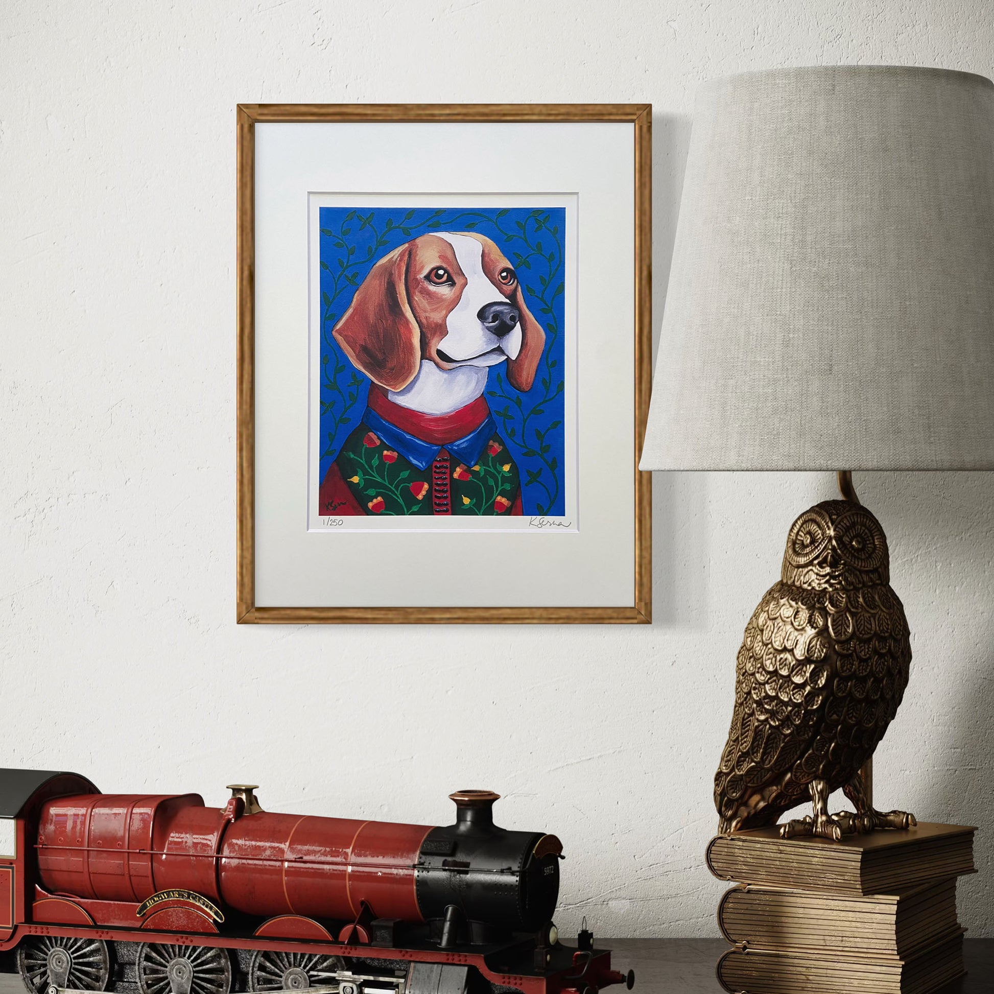 A framed print of Theo the Beagle hangs on a textured white wall, adding warmth and color to the space. The print is encased in a natural wood frame with a white mat. Below, a cozy vignette includes a decorative brass owl lamp sitting on stacked books and a model steam locomotive, creating a vintage-inspired aesthetic.