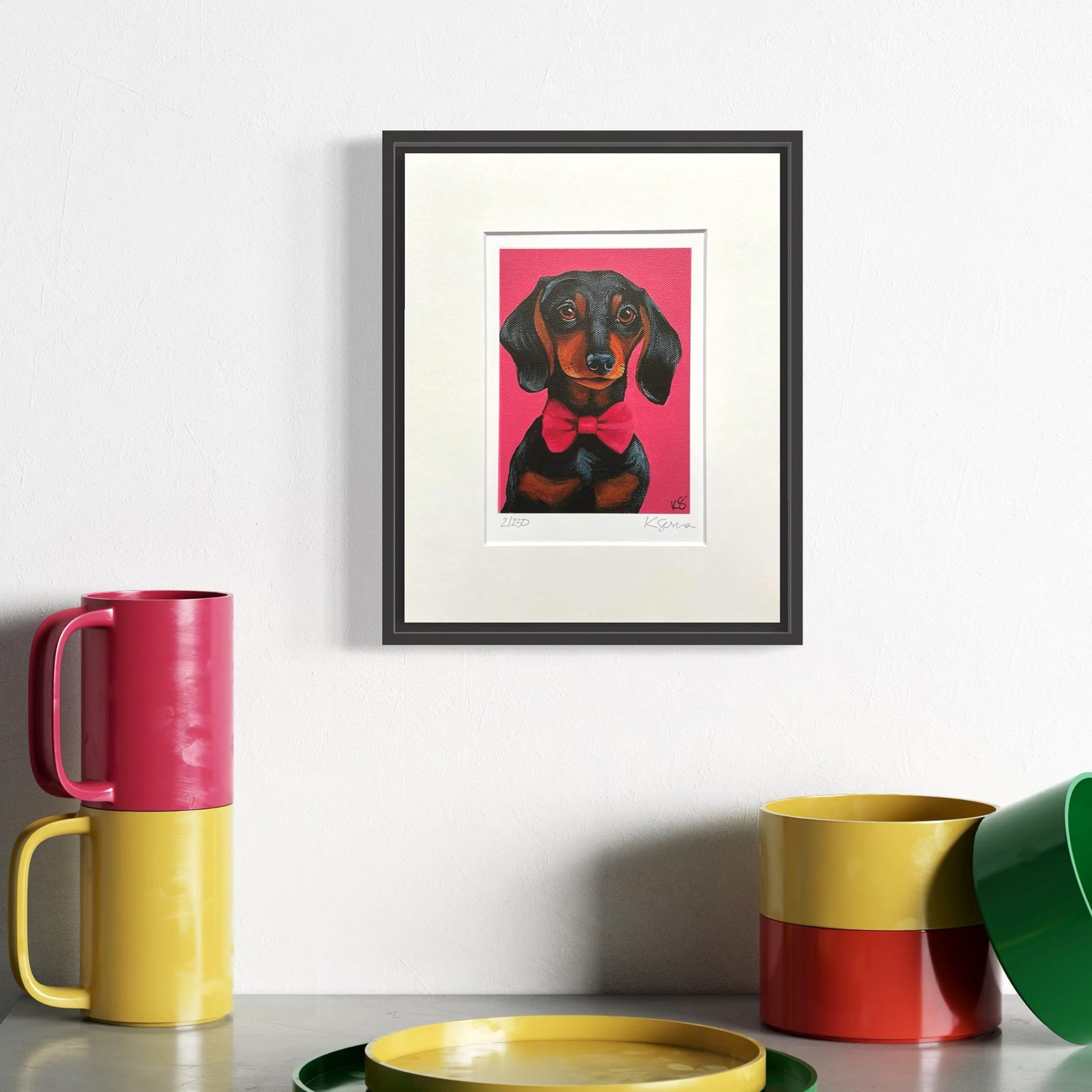 Framed artwork of a dog with a pink bow tie on a white wall, with colorful mugs and a green bowl in the foreground.