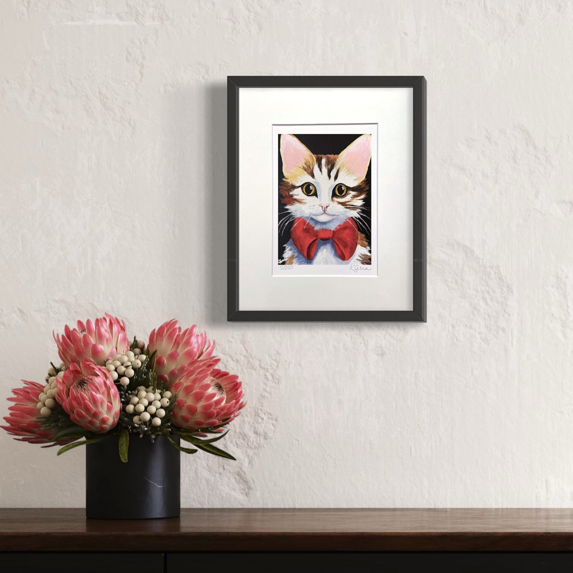 Print of brown and white cat with red bow, hanging on wall above a table with flowers