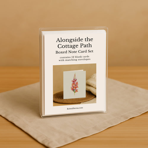 Boxed note card set titled 'Alongside the Cottage Path' on a beige surface with a brown background.