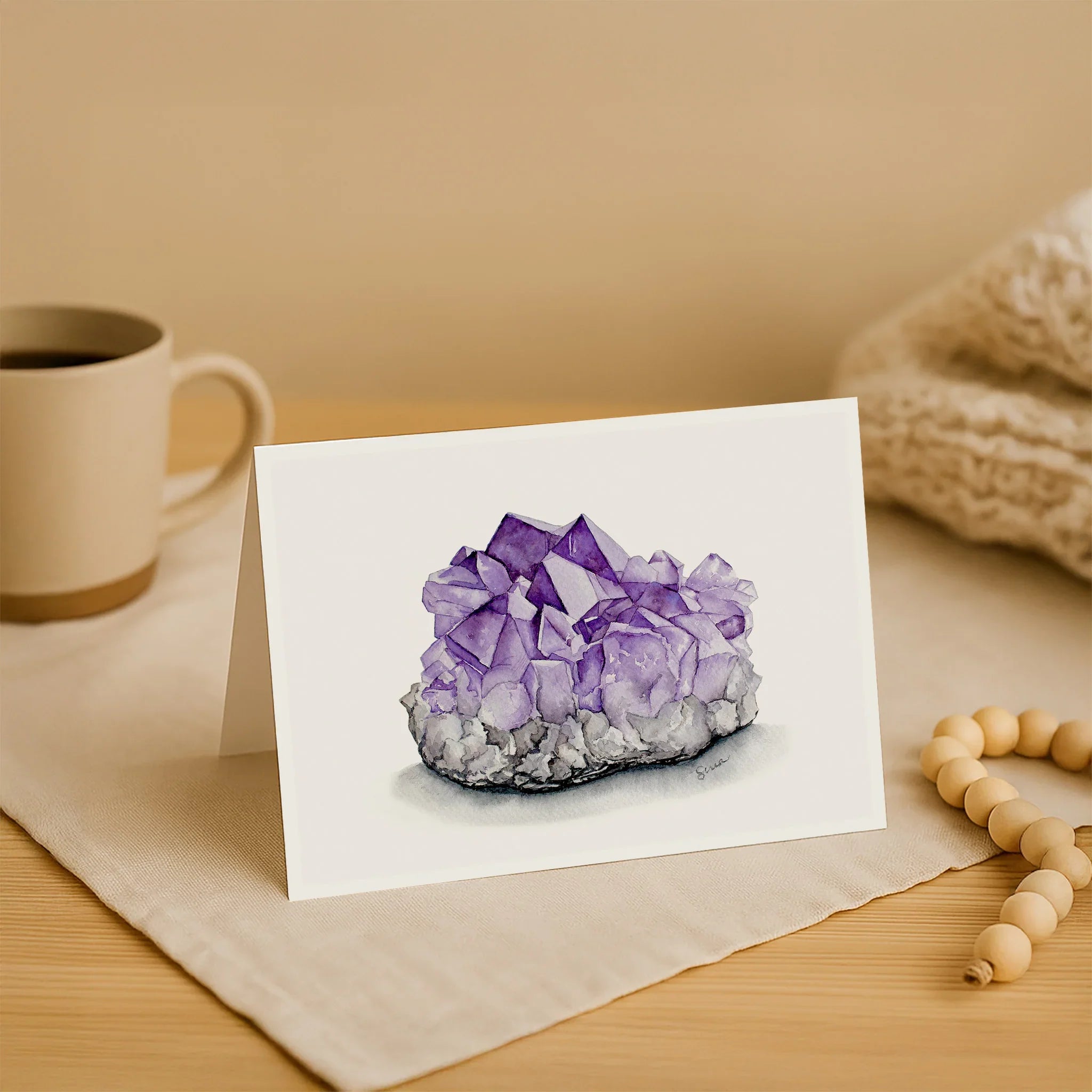 Note card with a purple crystal illustration on a wooden surface with a mug and beads.