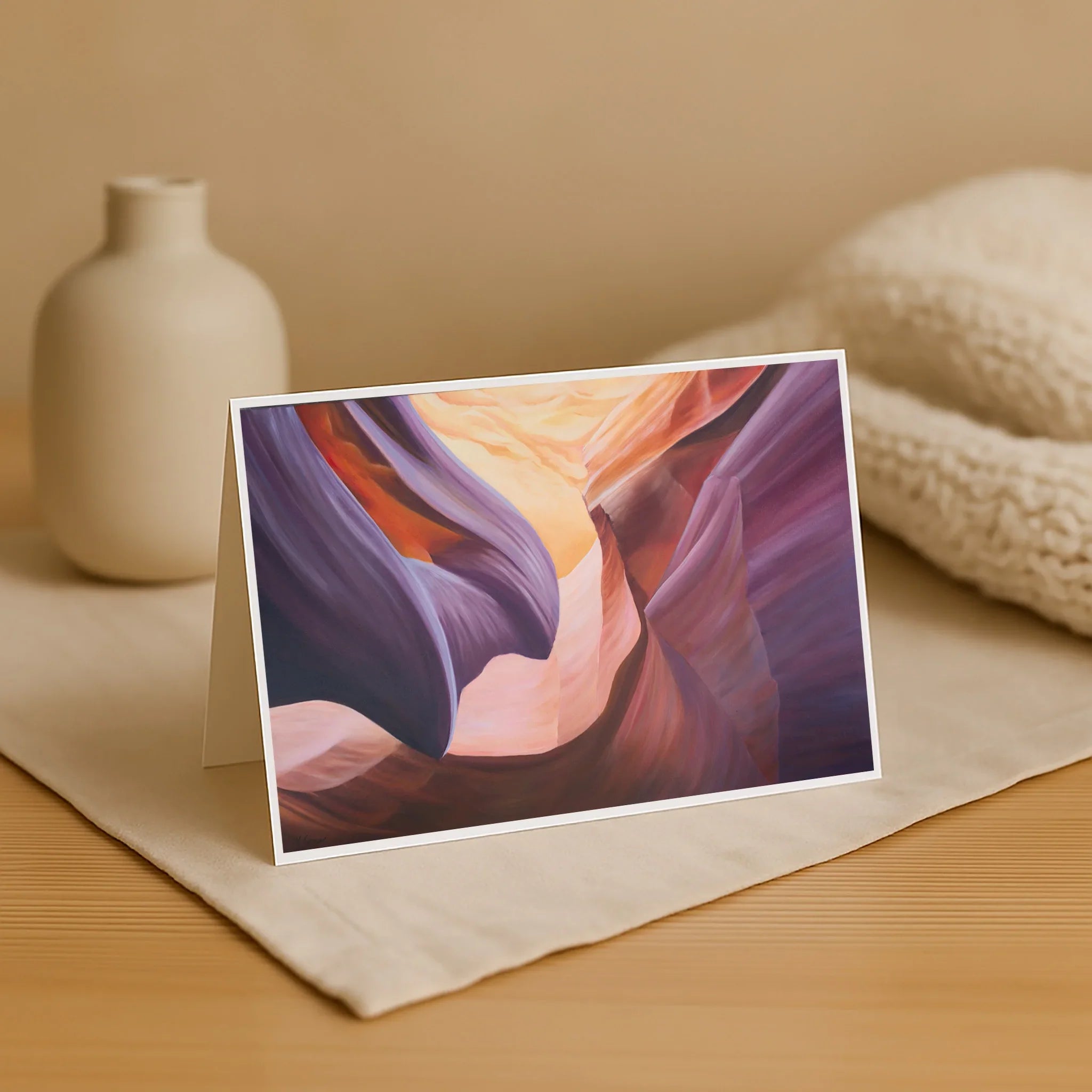 Greeting card with a colorful landscape design on a wooden surface with a vase and fabric in the background.