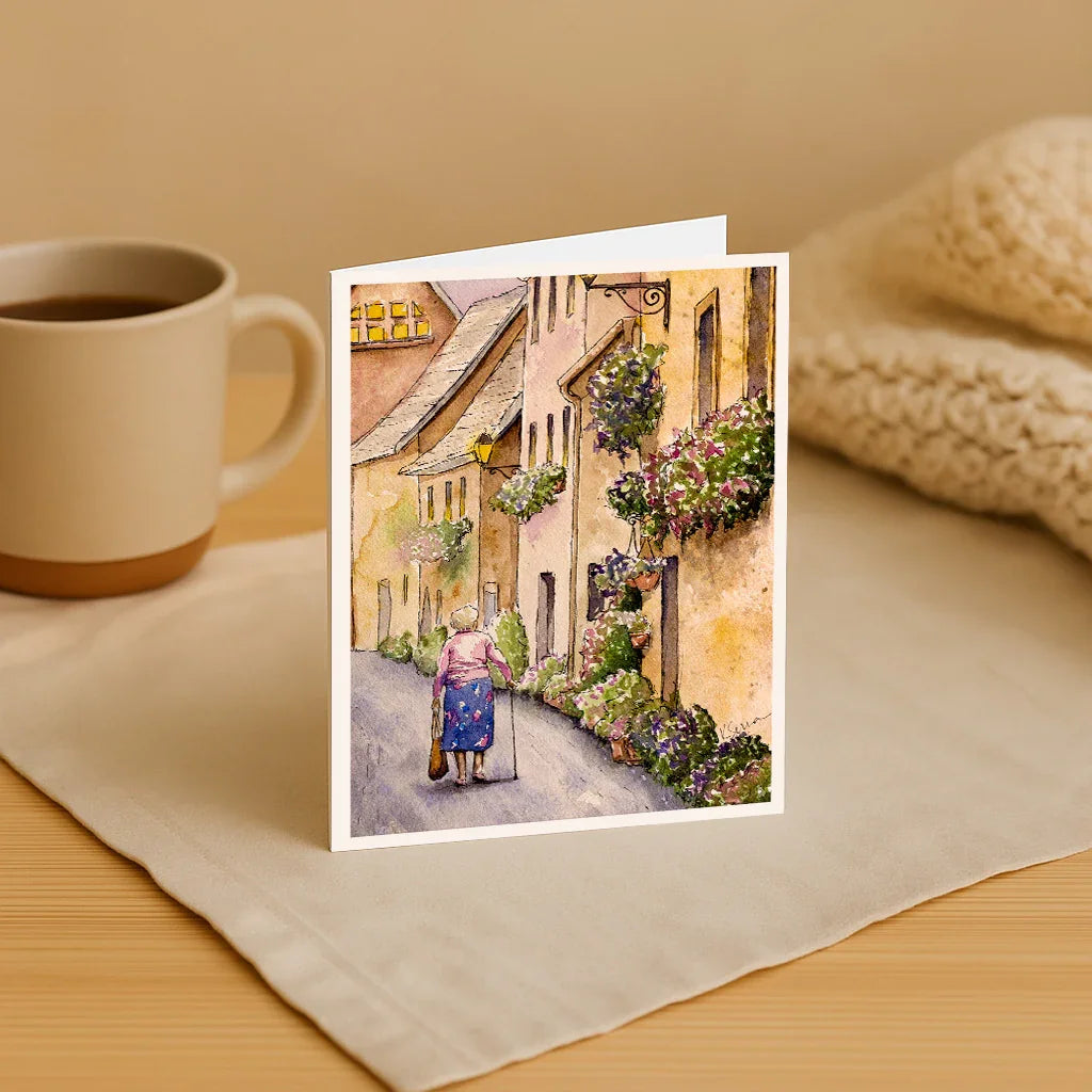 Greeting card with a painting of an elderly couple walking down a street, next to a cup of coffee on a wooden surface.