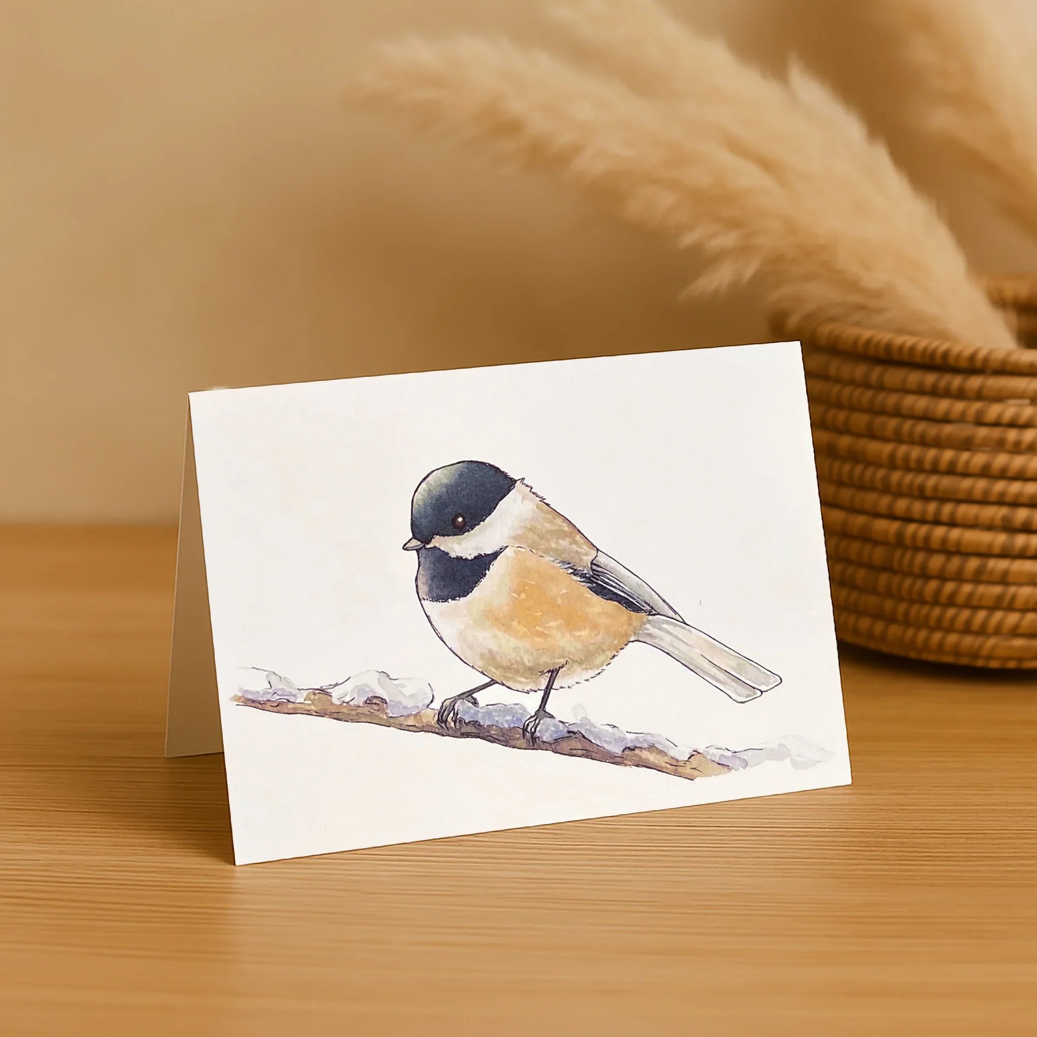 Greeting card with a chickadee illustration on a wooden surface with a basket in the background