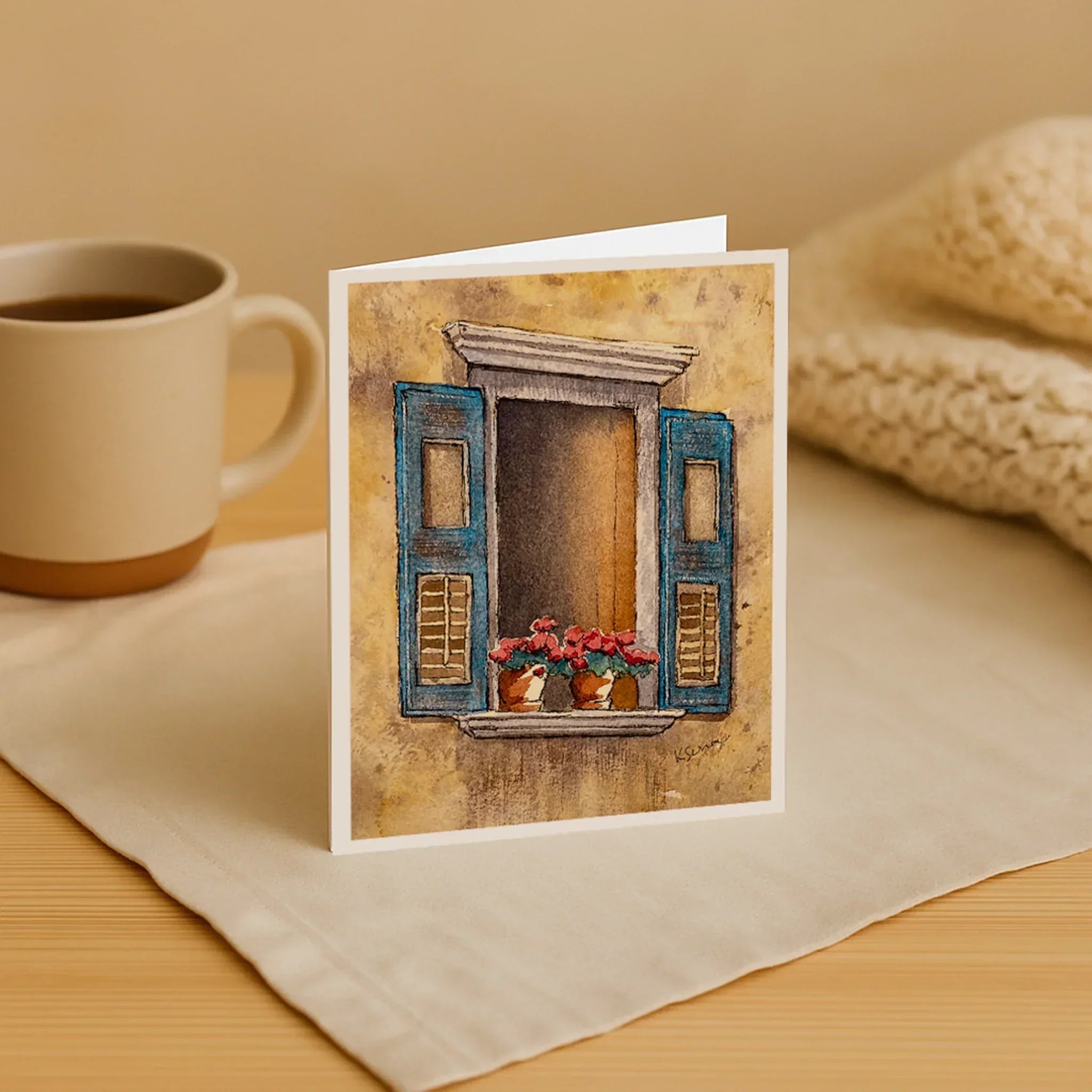 Greeting card with a window design on a table with a mug and blanket.