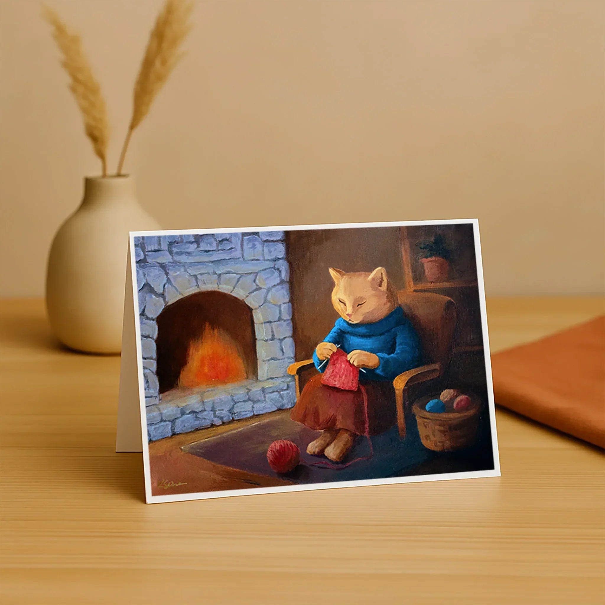 Greeting card with a cat knitting by a fireplace on a wooden surface.