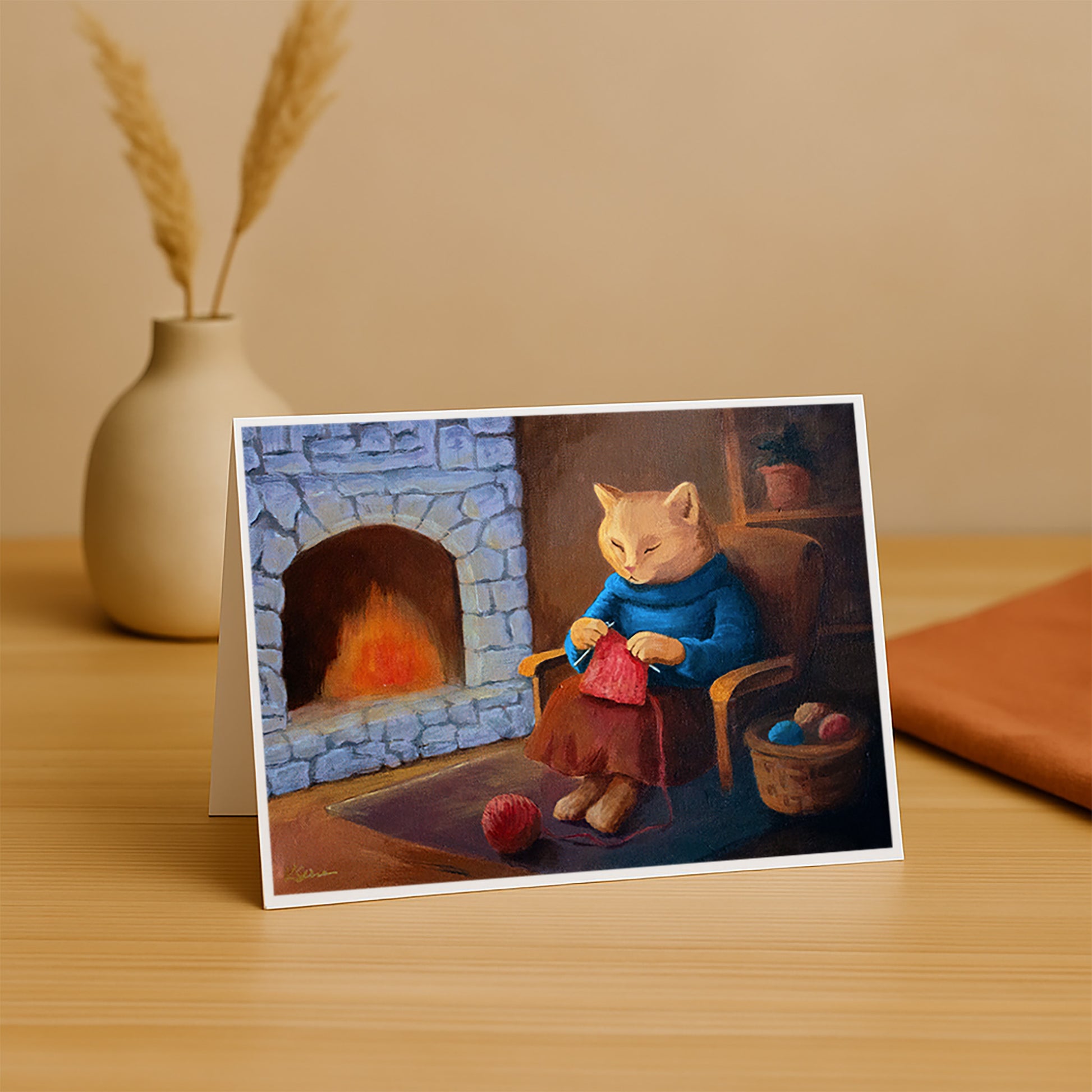 Greeting card with a cat knitting by a fireplace on a wooden surface.