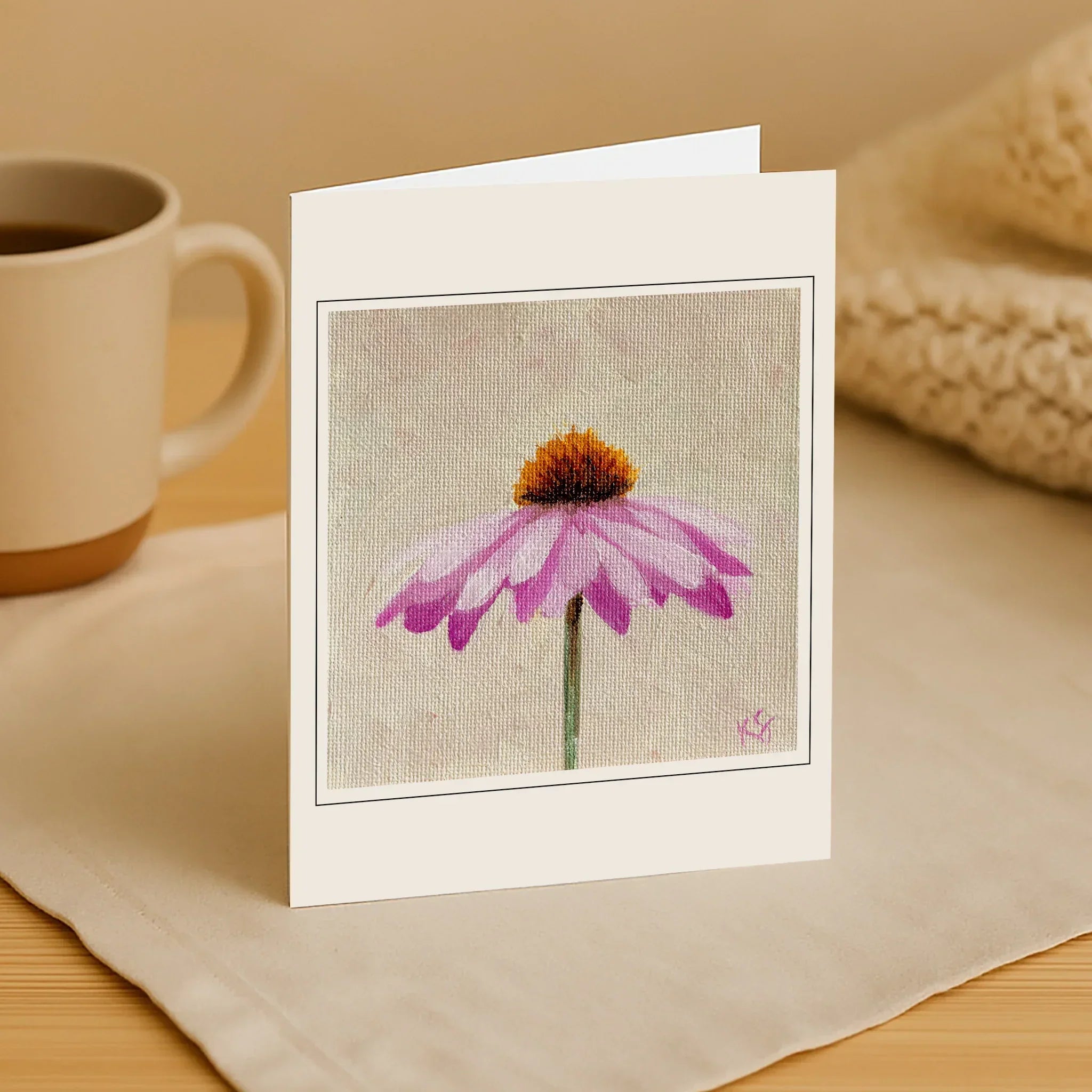 Greeting card with a purple flower design on a wooden surface with a mug and fabric.