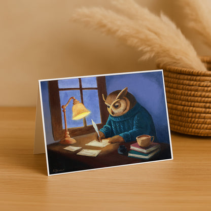 Greeting card with an owl in a blue sweater writing at a desk, placed on a wooden surface.