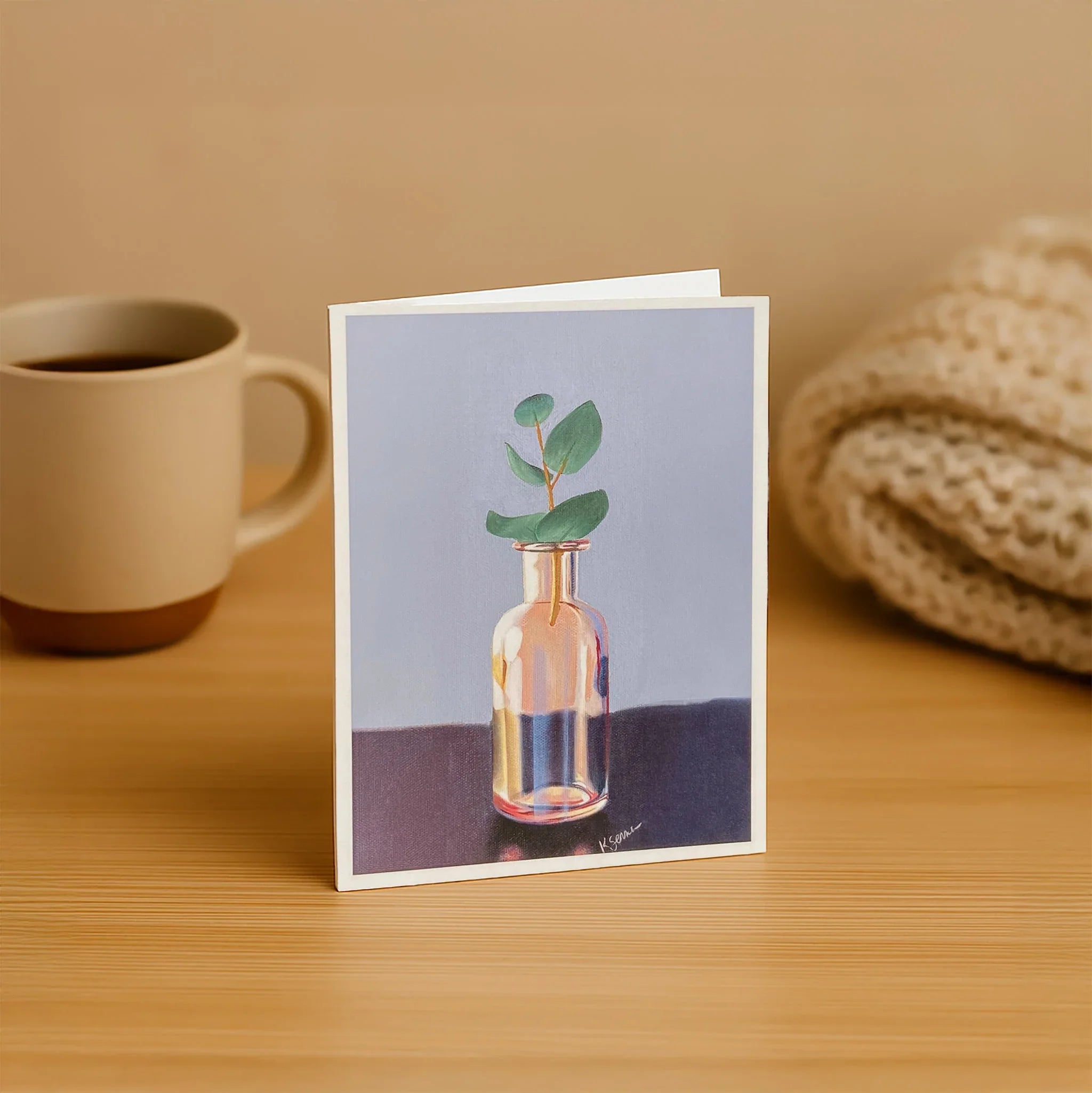 Greeting card with a bottle and plant design on a wooden table with a mug and knitted item in the background.