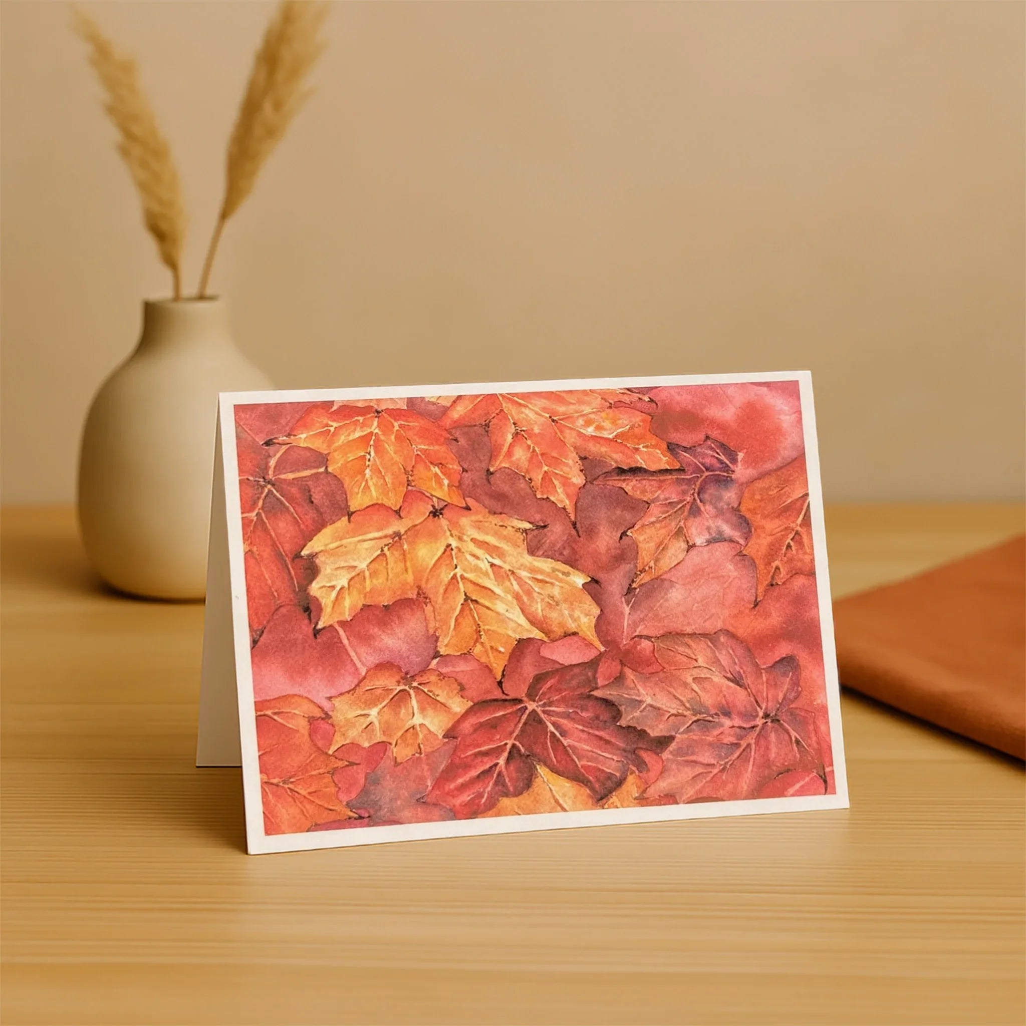 Greeting card with autumn leaf design on a wooden surface with a vase and book in the background.