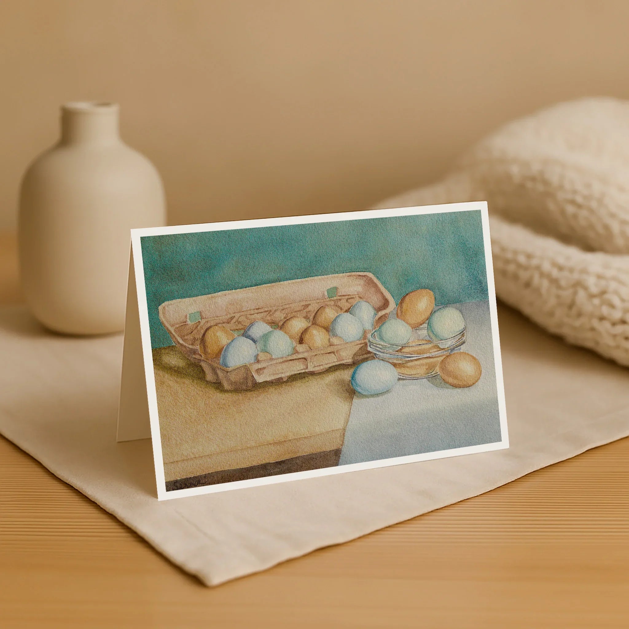 Greeting card with a design of eggs in a carton on a beige surface with a vase and woven item in the background.
