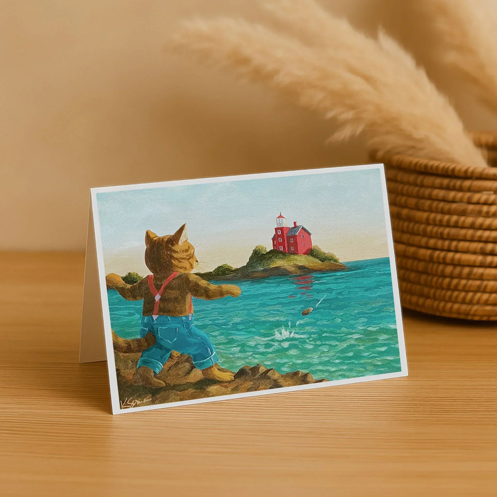 Greeting card with a cat character by the sea, featuring a lighthouse, on a wooden surface.