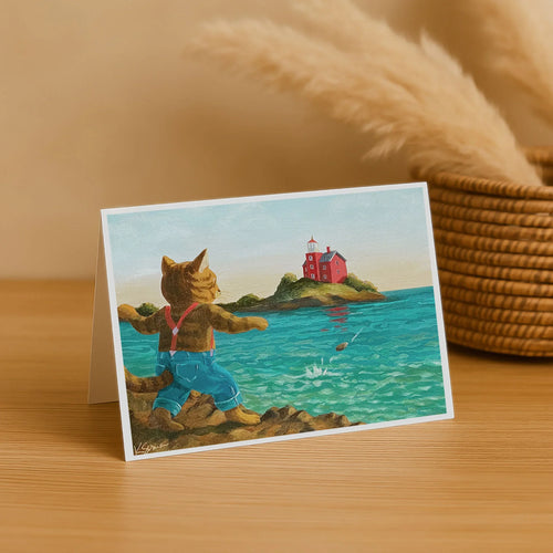 Greeting card with a cat character by the sea, featuring a lighthouse, on a wooden surface.