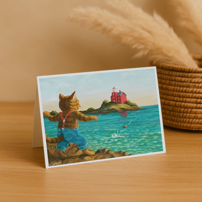 Greeting card with a cat character by the sea, featuring a lighthouse, on a wooden surface.