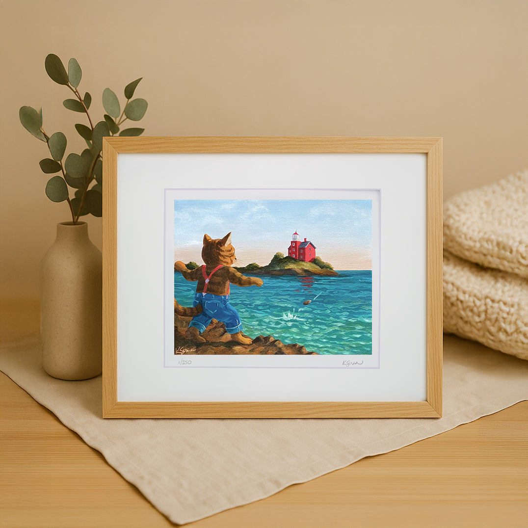 Framed artwork of a cat by the sea with a lighthouse, placed on a wooden surface.