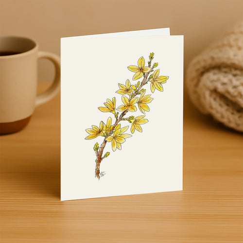 Greeting card with yellow floral design on a wooden surface with a mug and fabric in the background.