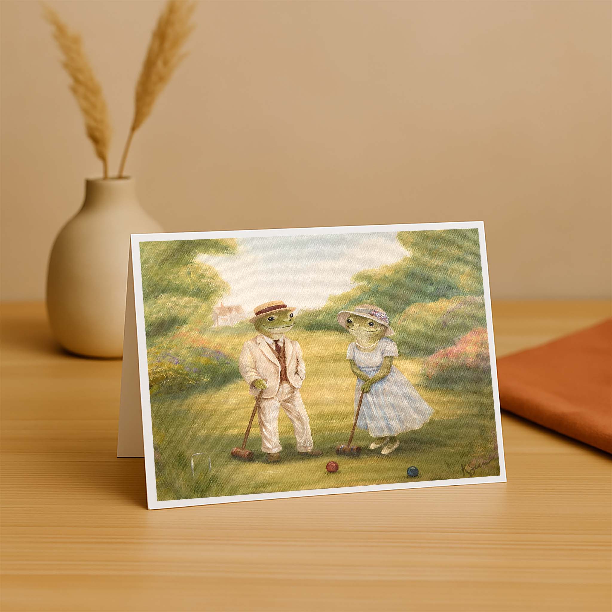 Greeting card with illustration of two frogs playing croquet on a wooden surface.