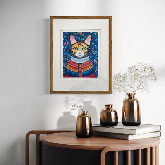 A framed print of Hazel the Cat hangs on a light-colored wall, adding a touch of warmth and vibrancy to the space. The artwork is encased in a natural wood frame with a white mat. Below, a modern wooden console holds a trio of metallic bronze vases with delicate white flowers, along with stacked books, creating an elegant and artistic display.