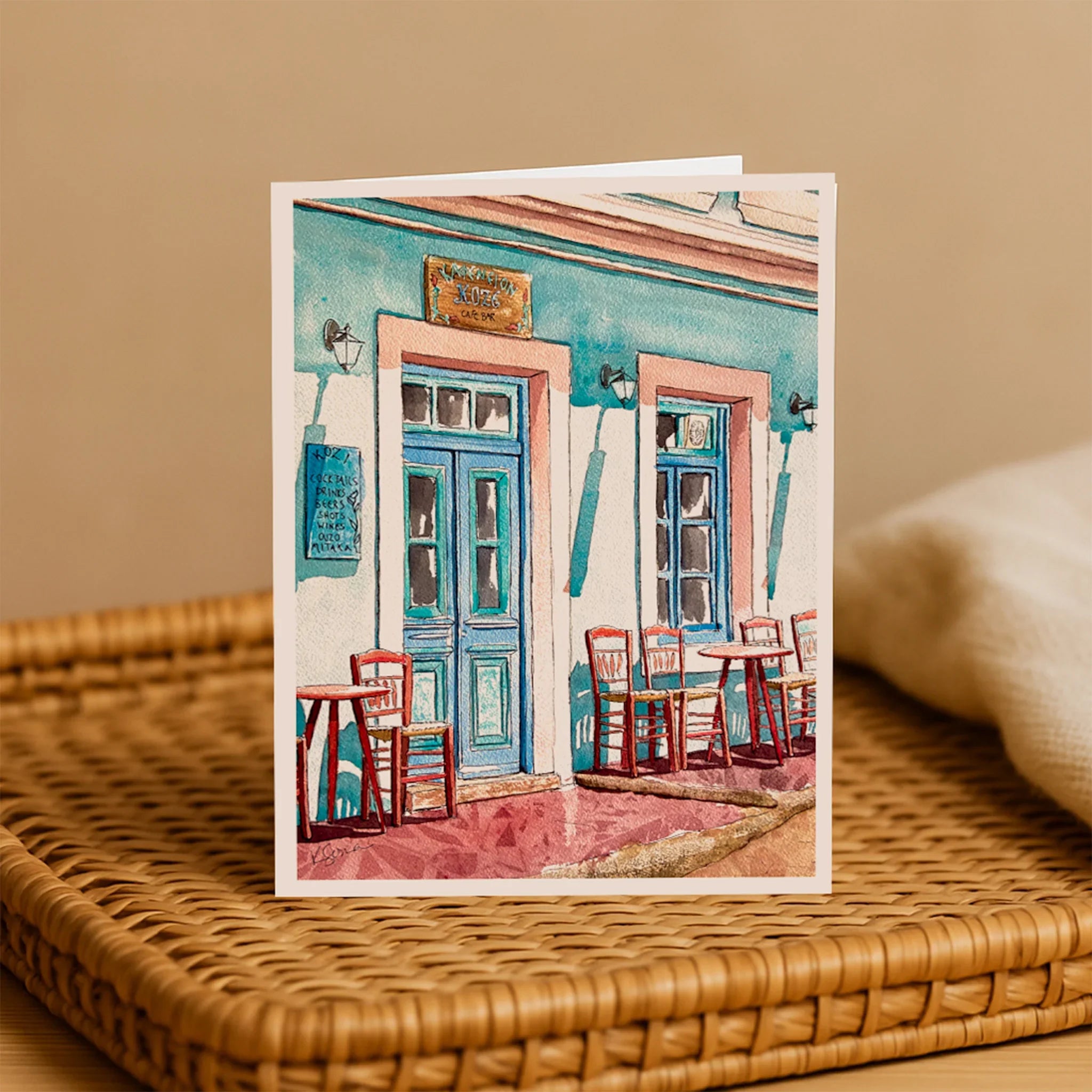 Greeting card with a painting of a colorful building and outdoor seating on a woven surface.