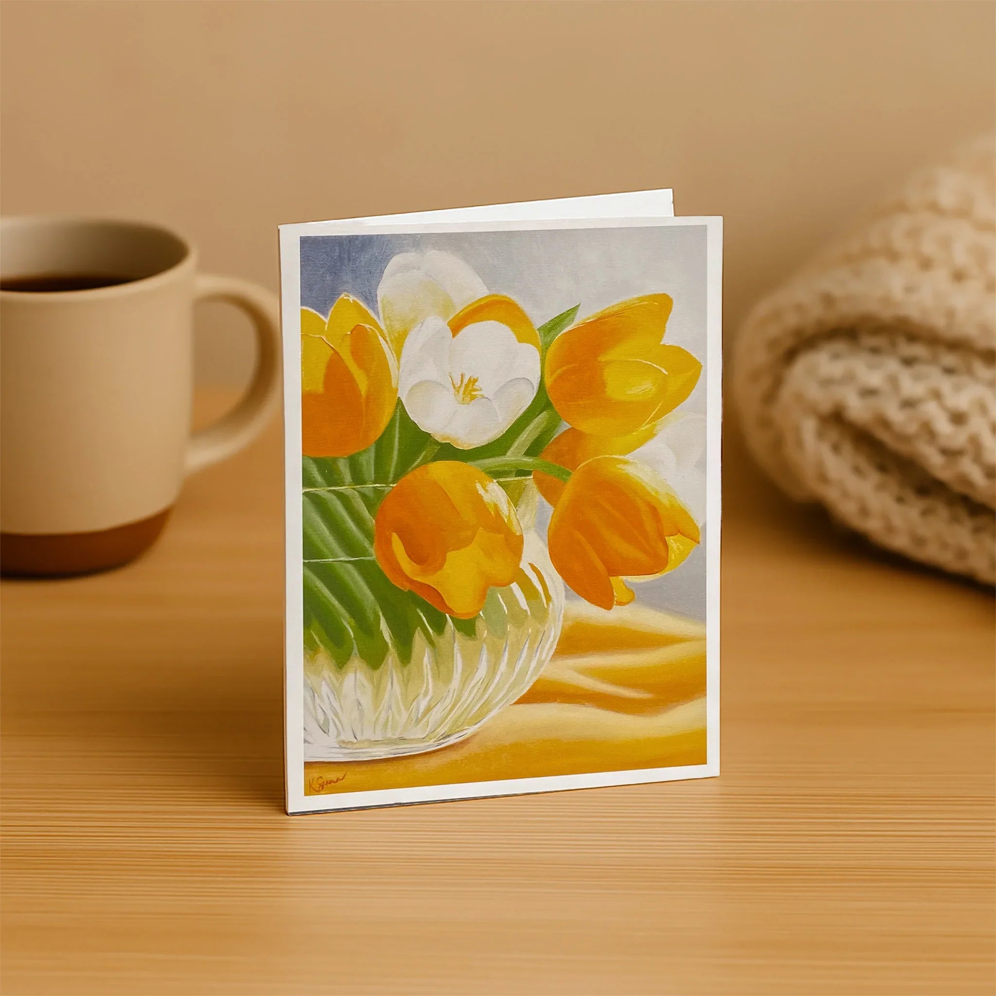 Greeting card with a floral design on a wooden surface next to a mug and knitted item.