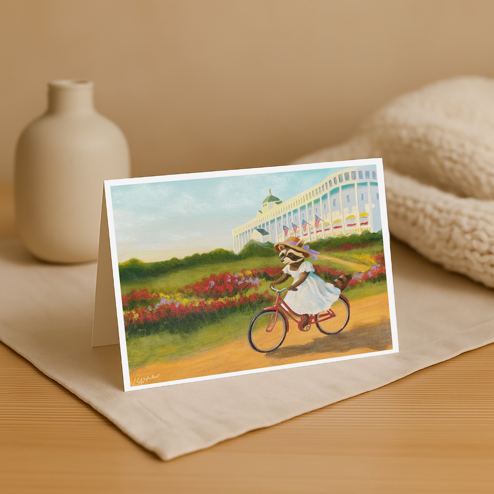 Greeting card with a raccoon riding a bike in a garden scene, placed on a beige surface with a vase and fabric in the background.