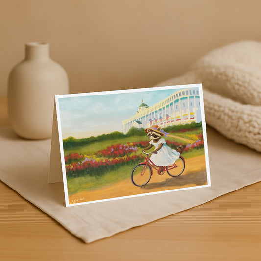 Greeting card with a raccoon riding a bike in a garden scene, placed on a beige surface with a vase and fabric in the background.