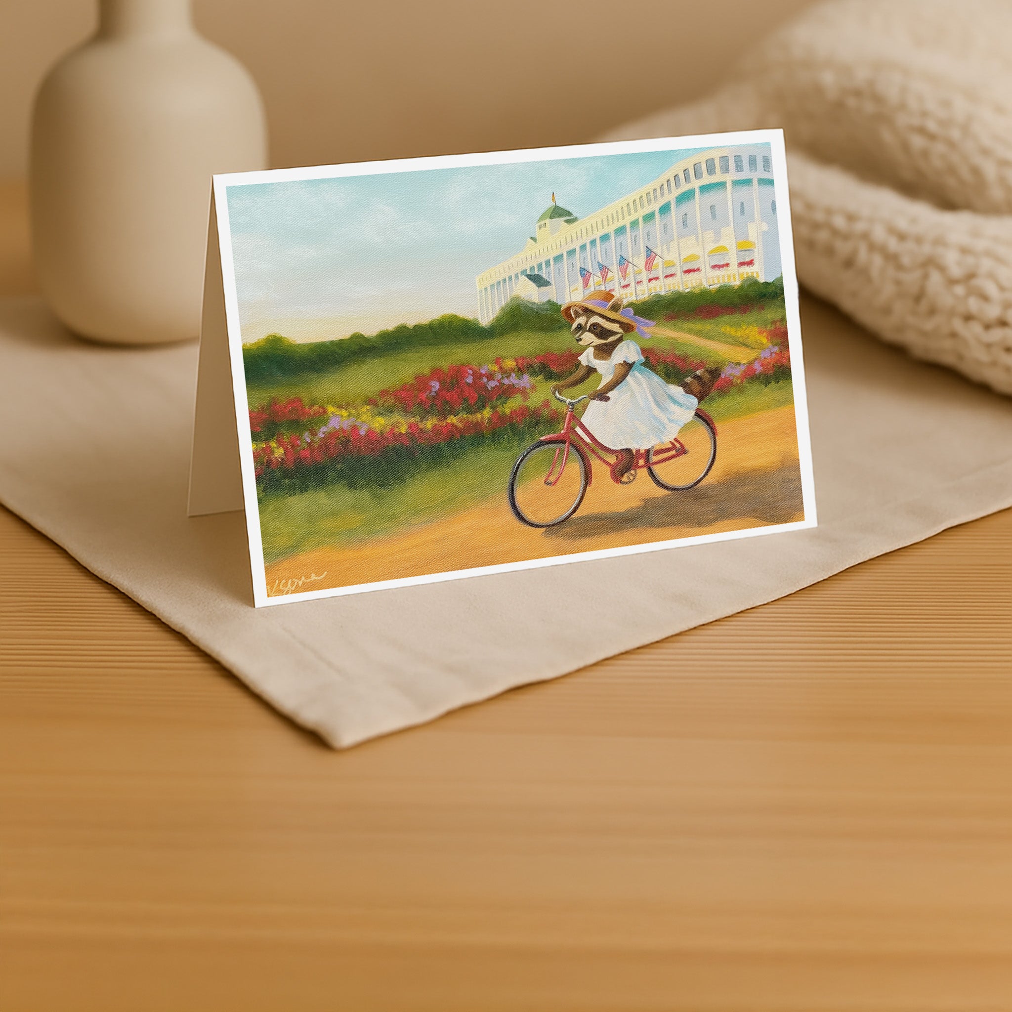 Greeting card with a raccoon on a bicycle in a garden setting, placed on a wooden surface.